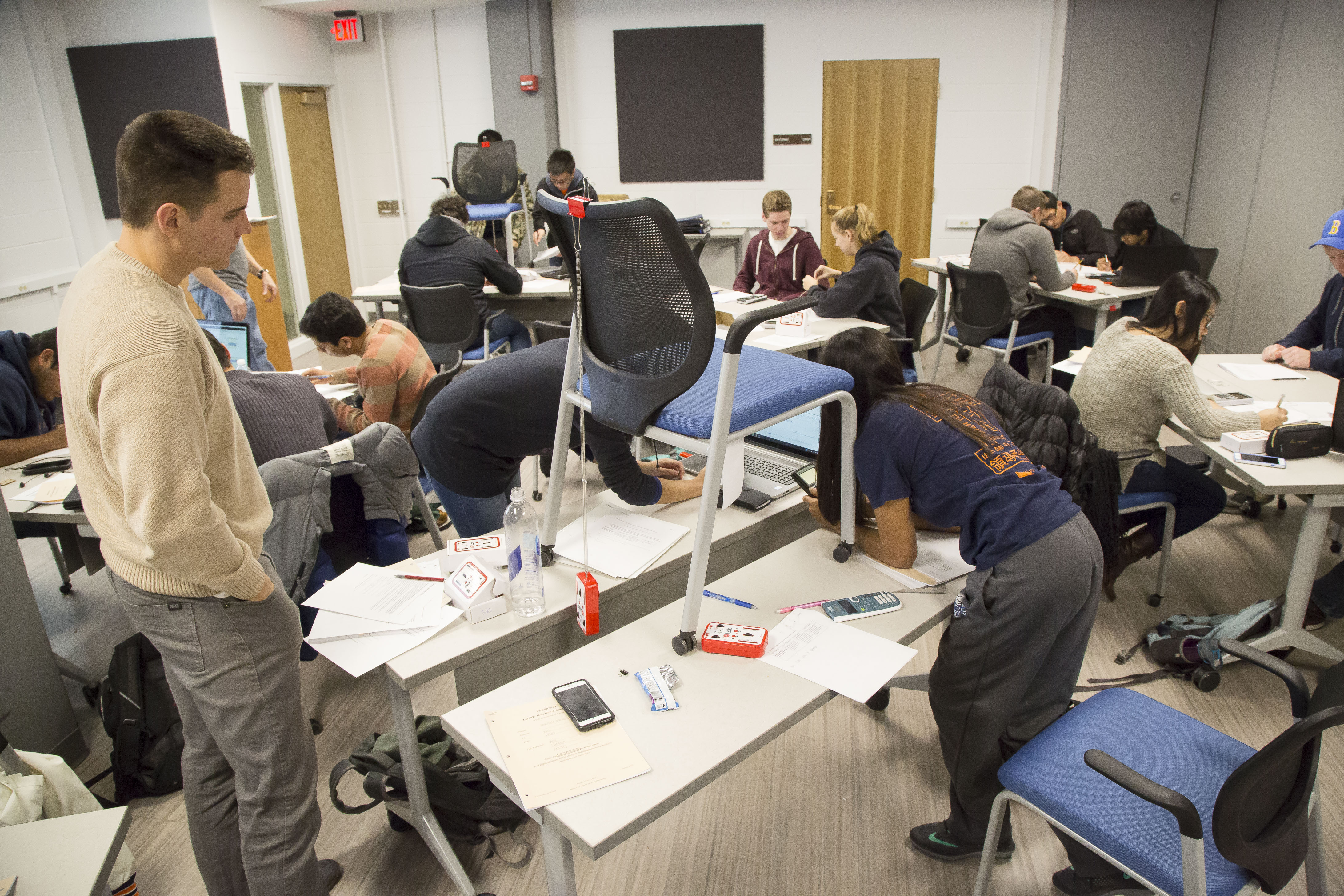 A photograph of a physics lab classroom. Some groups have placed chairs on top of their tables to hold iOLab devices attached to long springs.